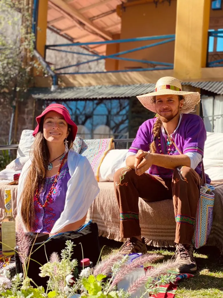 Una pareja con sombreros y ropa de colores sentada en el jardín, frente a la galería de la casa.