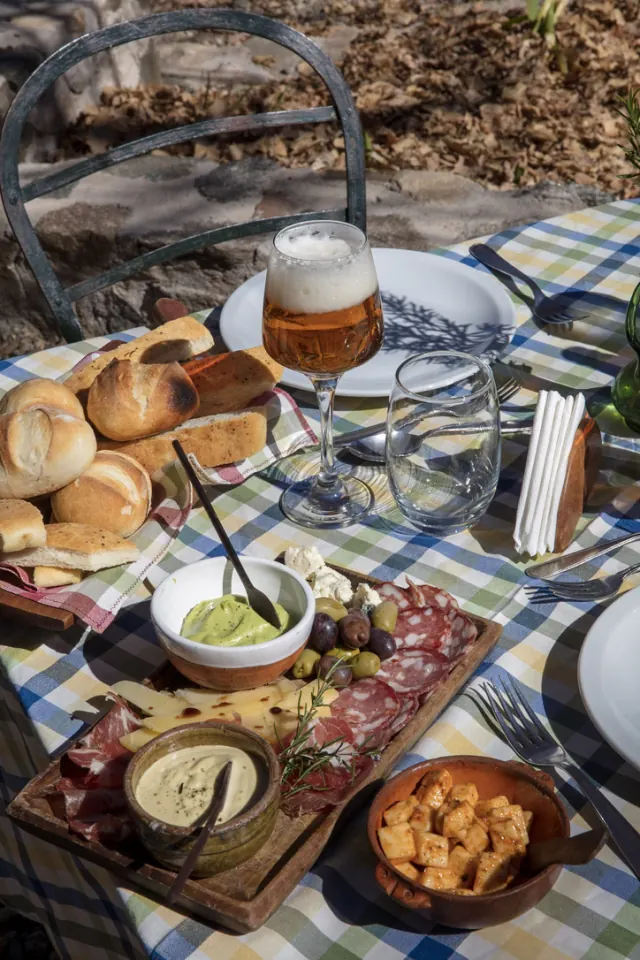 Una picada de fiambres, quesos, aceitunas y panes caseros sobre un mantel a cuadros, con una copa de cerveza, en la mesa de afuera.