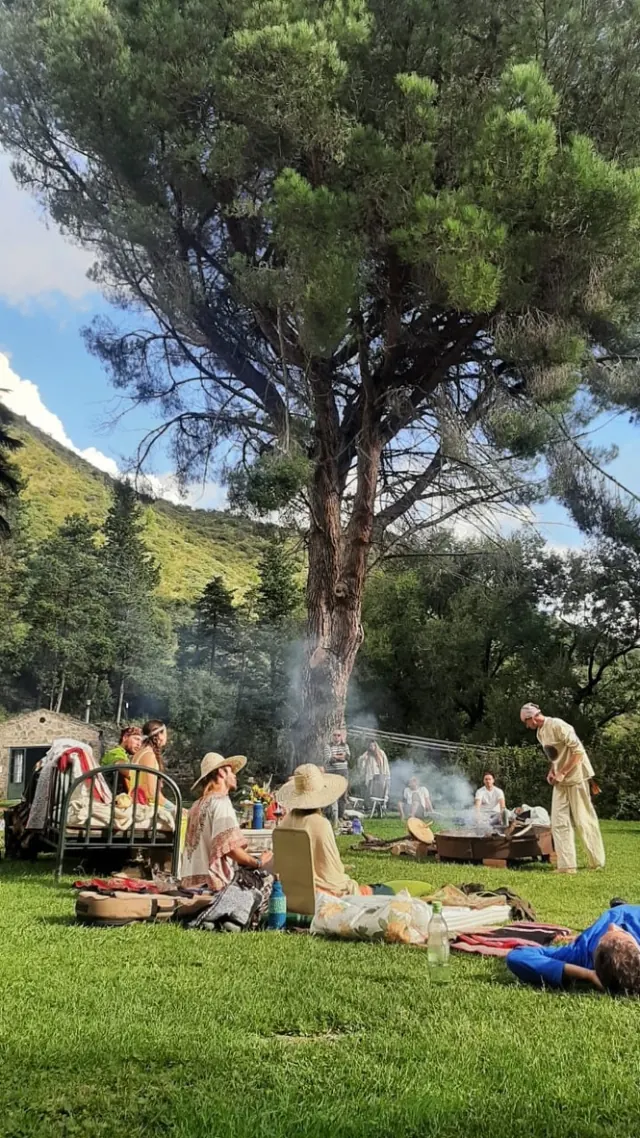 Un grupo con sombreros sentado en el pasto bajo el pino grande, con un fuego chico encendido y la sierra detrás.