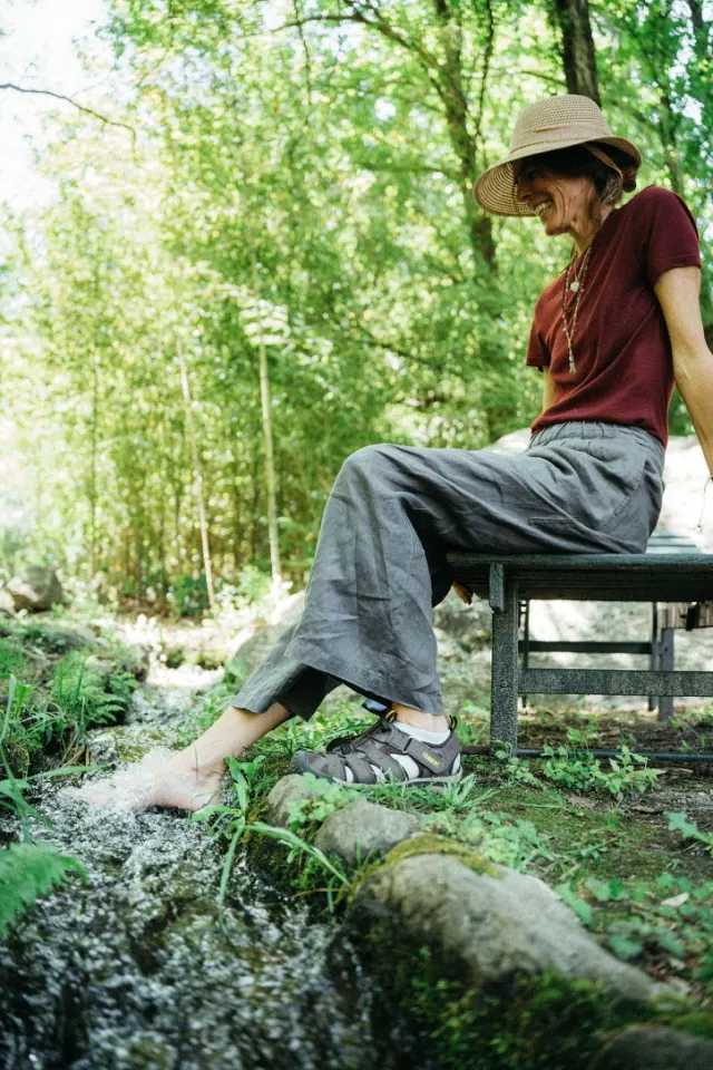 Una mujer con sombrero de paja sentada en un banco junto al arroyo, riéndose, con los pies en el agua.