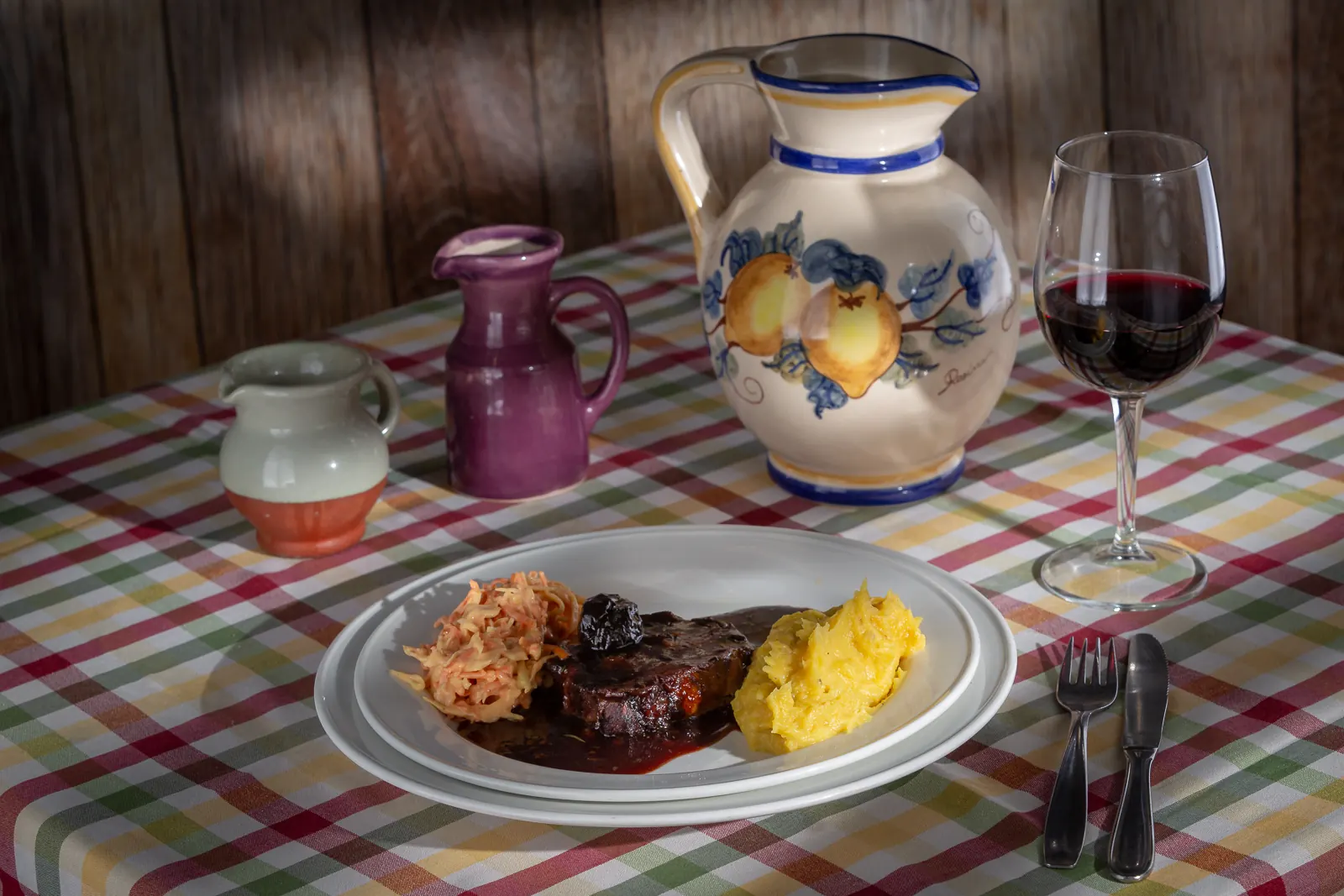 Un plato con carne braseada, puré de calabaza y ensalada de zanahoria, junto a una jarra de loza pintada y una copa de vino, sobre el mantel a cuadros.