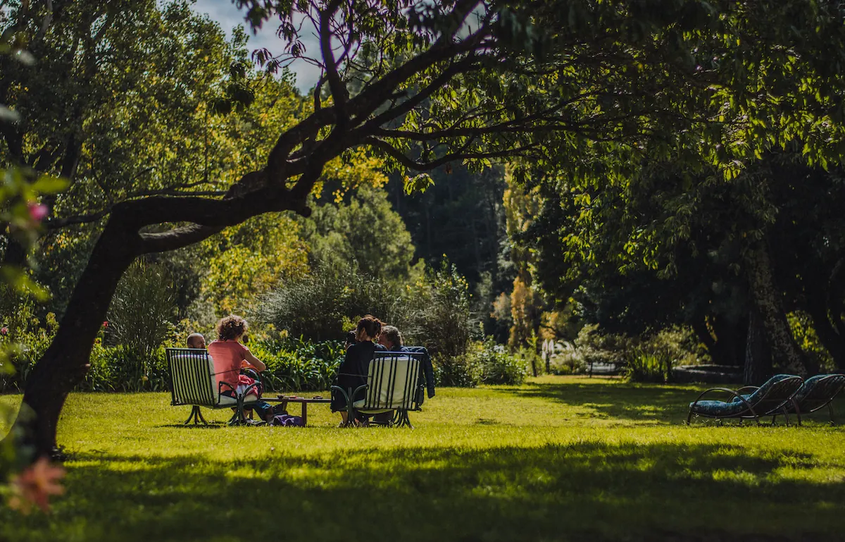 Dos personas conversando en reposeras en el medio del jardín, a la sombra de un árbol grande.