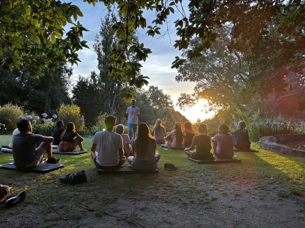 Un grupo sentado en colchonetas sobre el pasto escucha a un facilitador de pie, con el sol poniéndose entre los árboles del jardín.