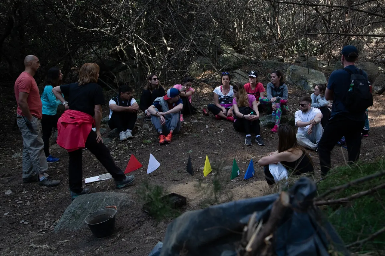 Un grupo sentado en ronda en el monte, a la sombra, frente a una fila de banderines de colores clavados en la tierra; una facilitadora habla de pie.
