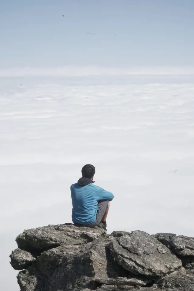 Un hombre de espaldas sentado en una roca del filo, mirando un mar de nubes que cubre el valle.