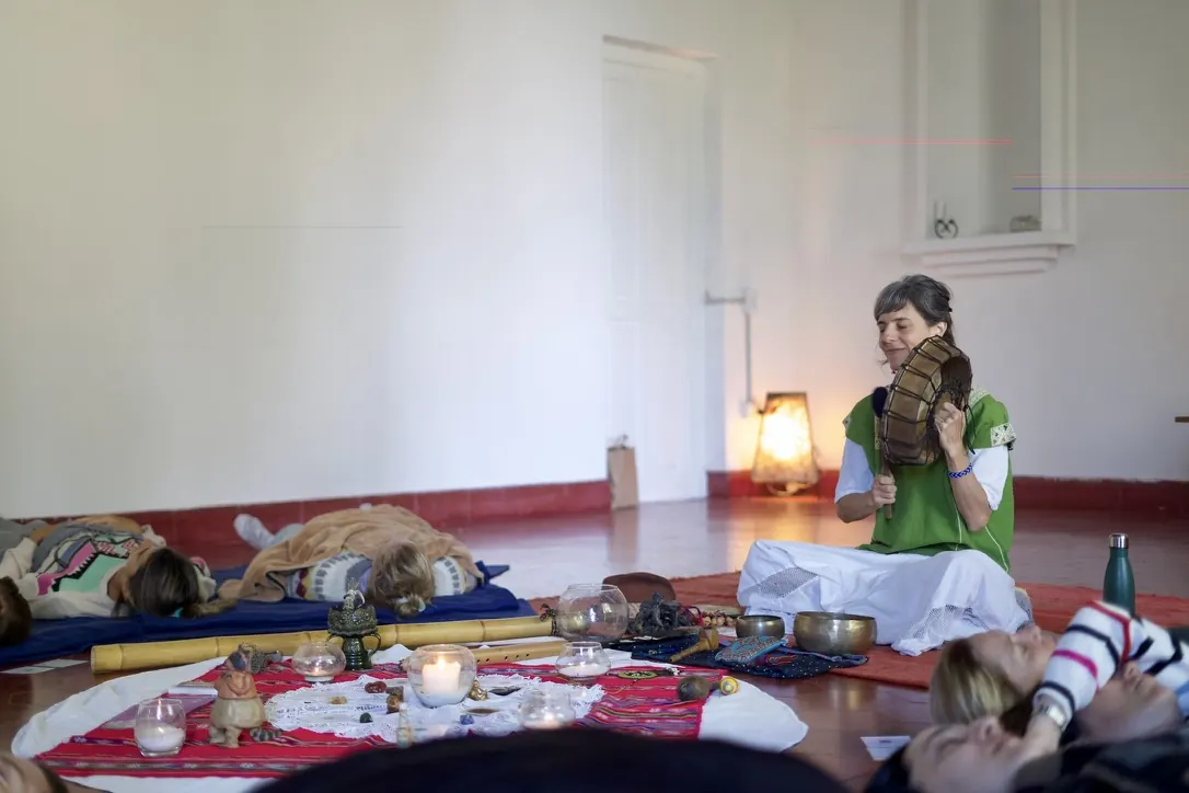 Una facilitadora sentada en el piso del salón toca un instrumento de sonido mientras el grupo descansa acostado en colchonetas, con cuencos y velas en el centro.