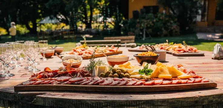 Una tabla larga de fiambres, quesos y aceitunas sobre una mesa de madera al aire libre, con copas y la casa al fondo.